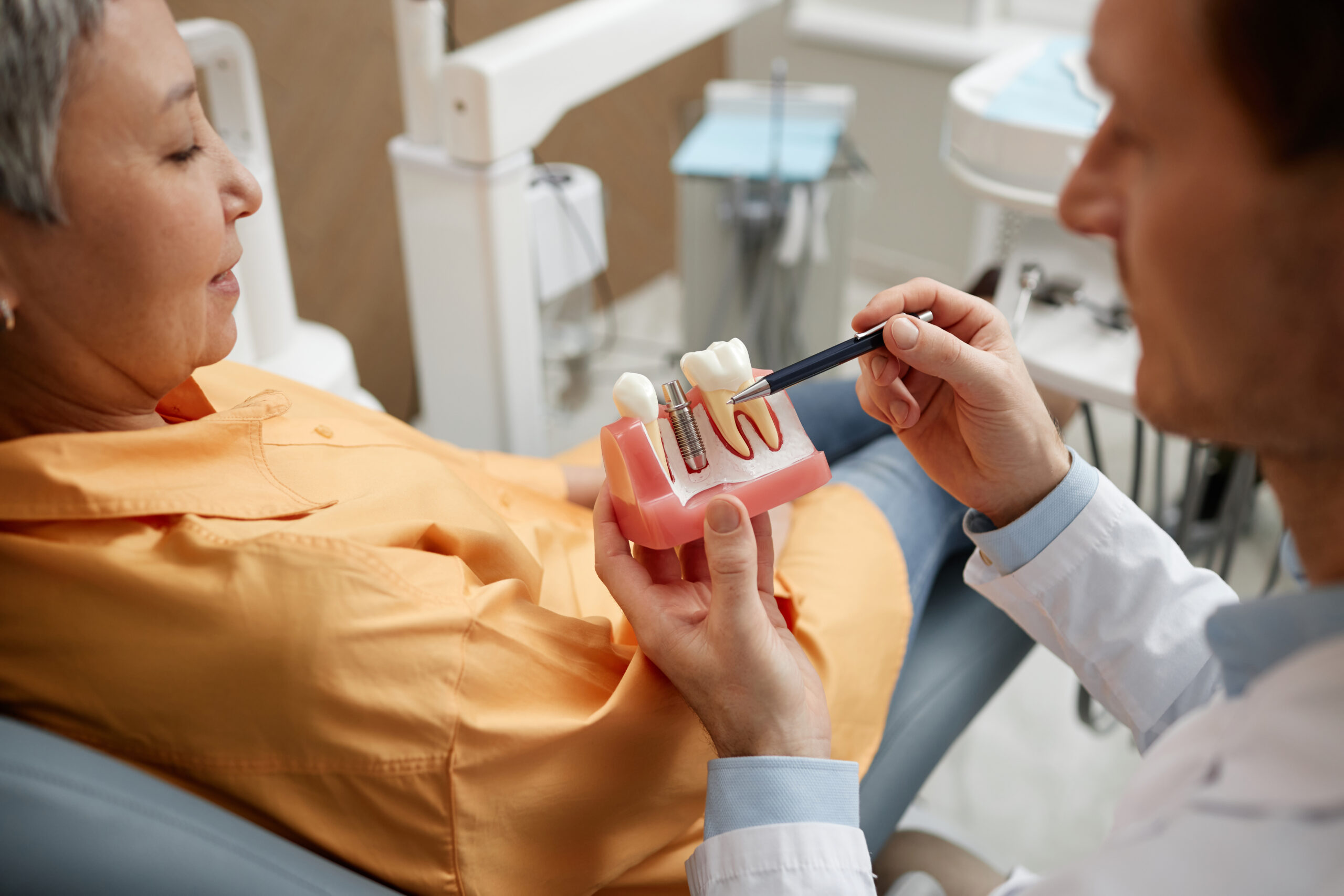 Dentist explaining a dental implant procedure to a seated patient using a model of teeth and gums in a dental office setting.