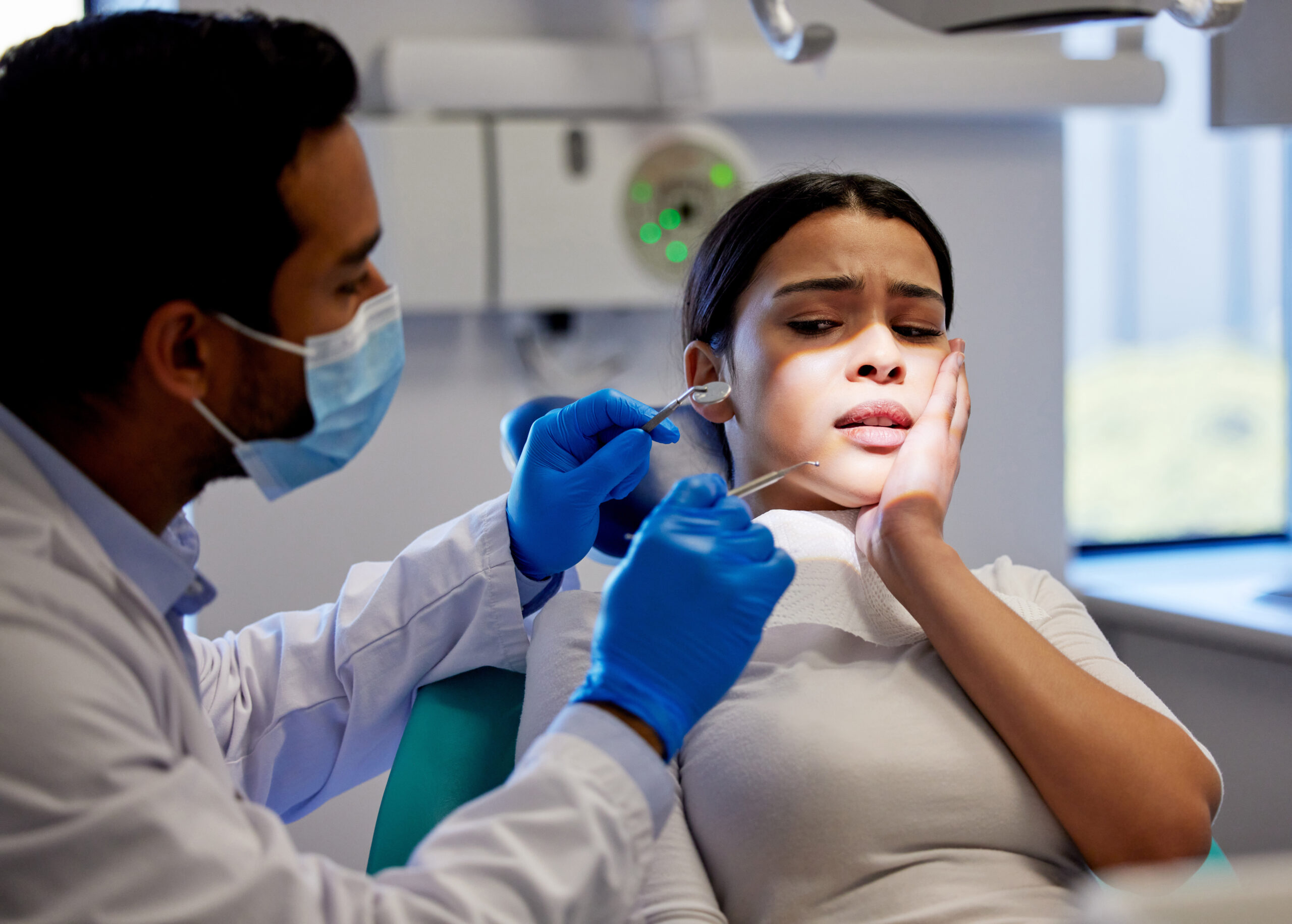 A concerned young woman sits in a dental chair holding her cheek in pain while a male dentist wearing a mask and blue gloves examines her mouth with dental tools