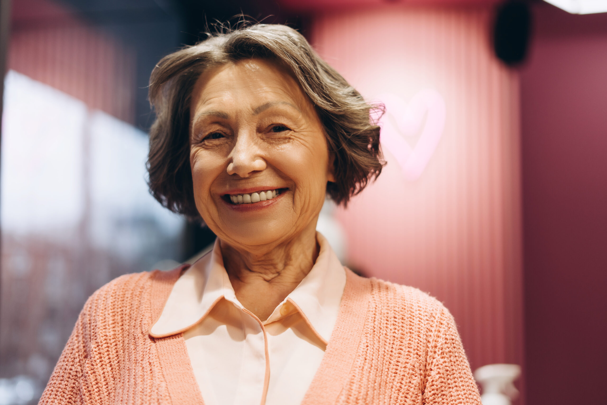 Smiling elderly woman wearing a peach cardigan and light blouse, standing indoors with a pink heart-shaped light in the background.