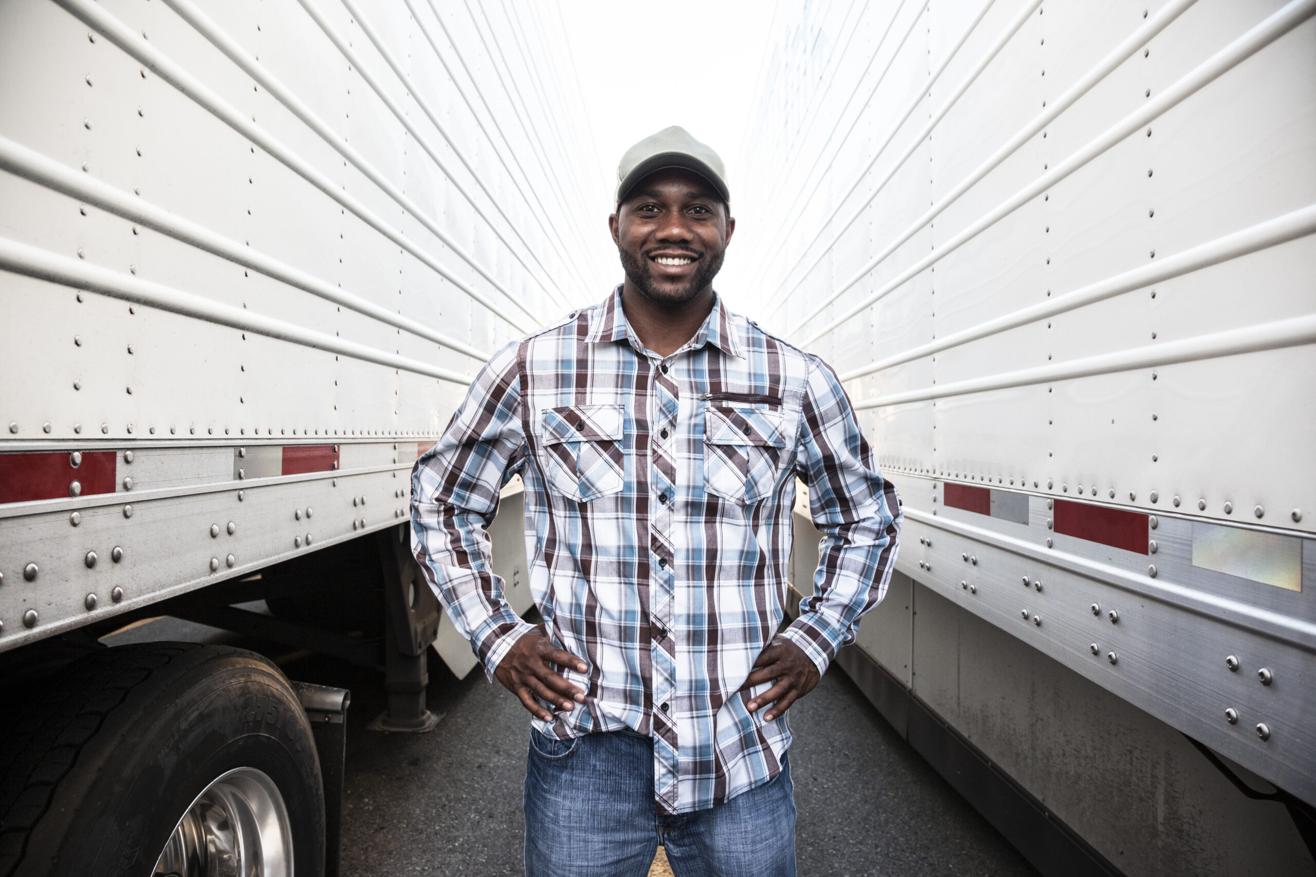 Smiling truck driver wearing a plaid shirt and cap standing confidently between two large white semi-trailers.